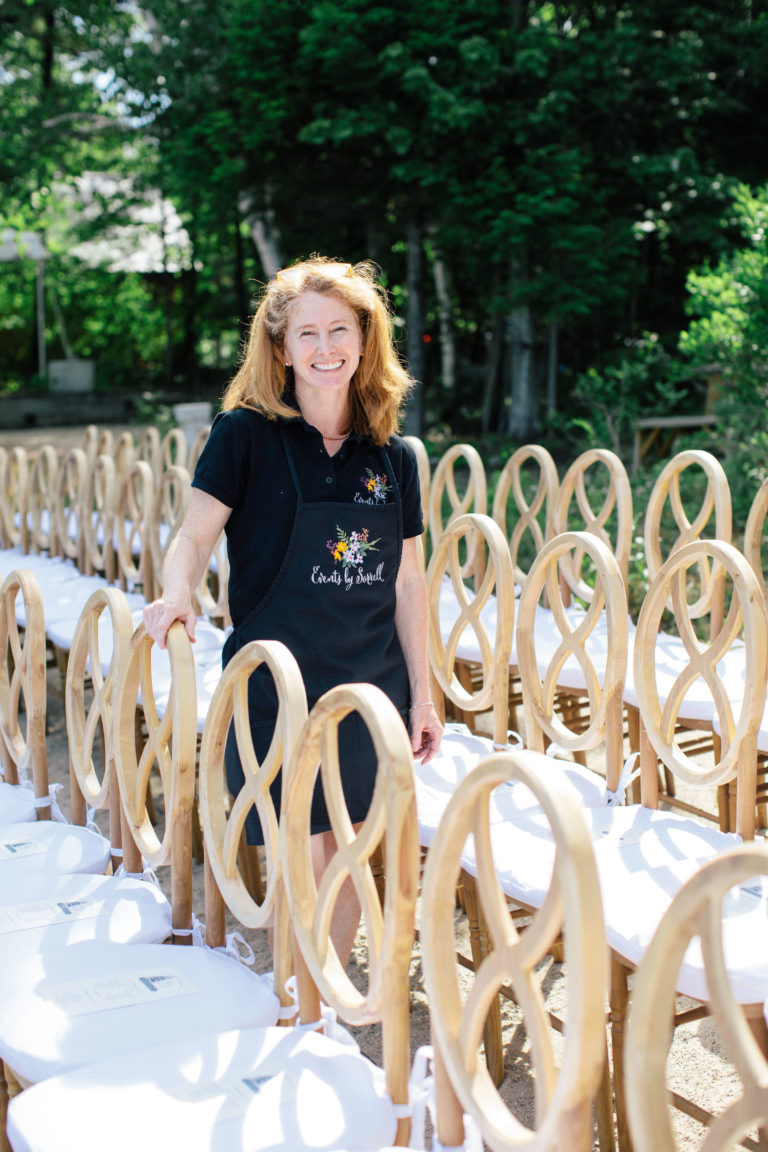 A woman stands among rows of wooden chairs outdoors.